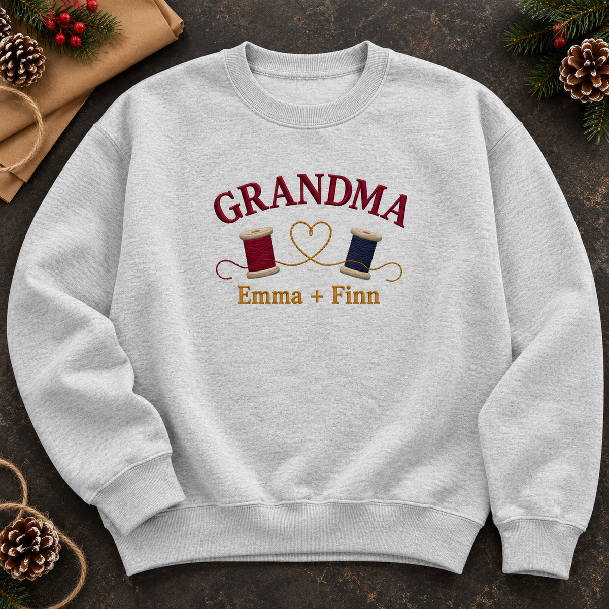 Grandma's Thread Spools sweatshirt concept mockup featuring two thread spools connected by a heart-shaped thread, sample personalization Emma + Finn, hero view