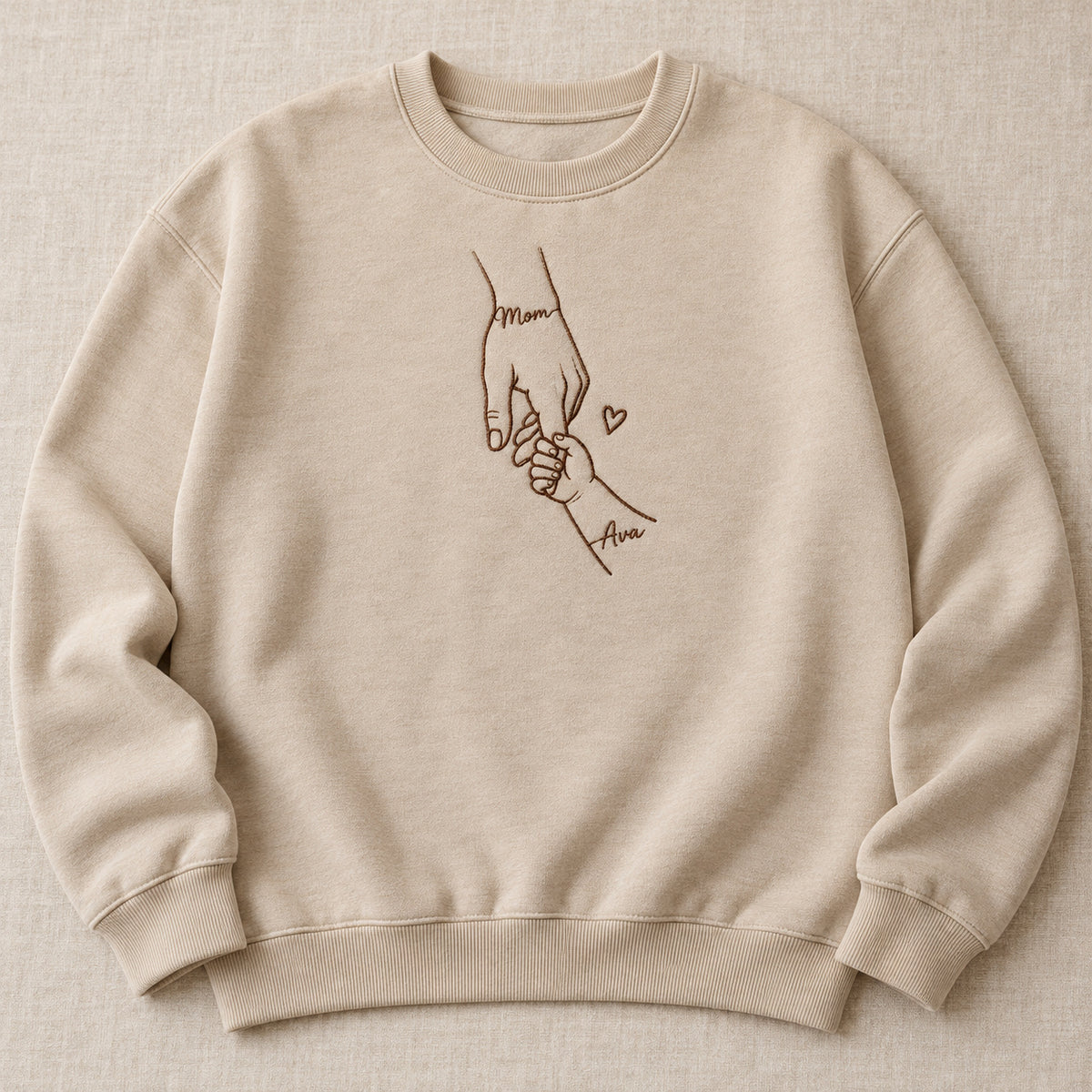 Sand adult sweatshirt mockup with centered mother-and-child hands embroidery and Mom / Ava lettering