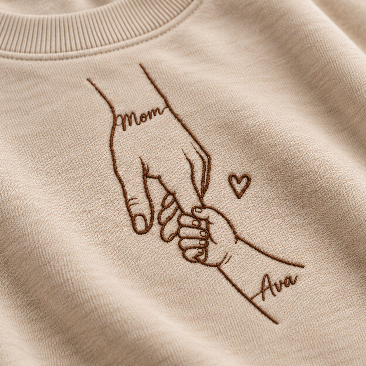 Close-up mockup of deep brown hands embroidery, small heart, and Mom / Ava lettering