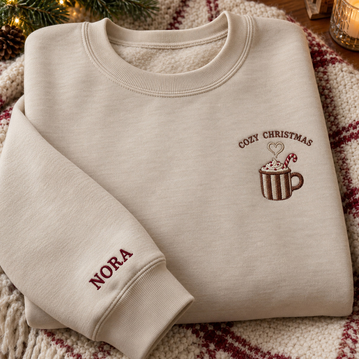 Cozy Christmas Cocoa sweatshirt concept mockup featuring a striped cocoa mug with a candy cane and heart-shaped steam, sample personalization NORA, hero view