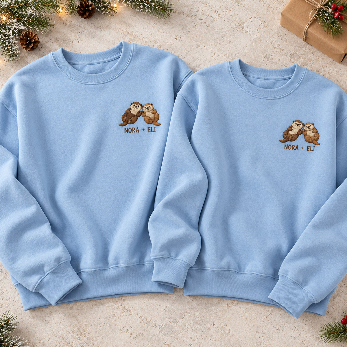 Holding Hands Otters sweatshirt concept mockup featuring two otters holding paws, sample personalization NORA + ELI, hero view
