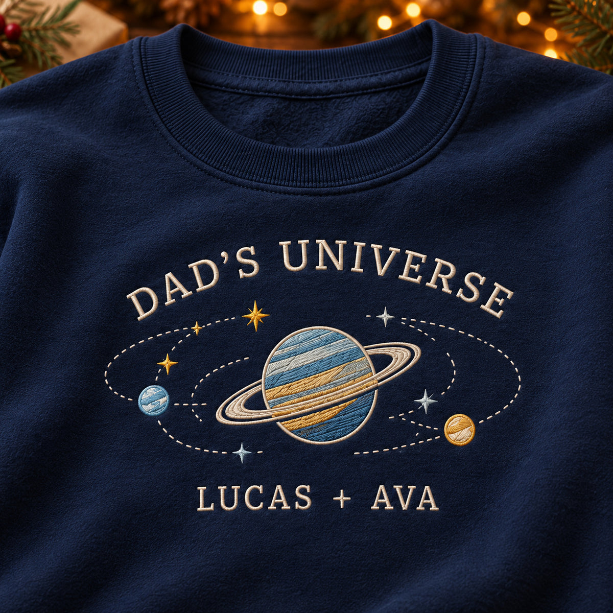 Dad's Universe sweatshirt concept mockup featuring a ringed planet with smaller planets and stars, sample personalization LUCAS + AVA, embroidery view