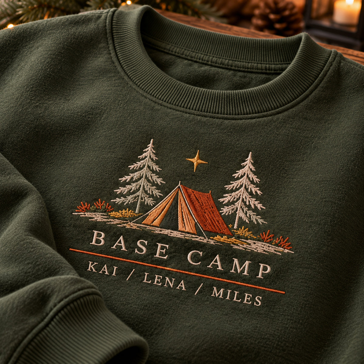 Family Base Camp sweatshirt concept mockup featuring a camping tent between evergreen trees beneath a star, sample personalization KAI / LENA / MILES, embroidery view