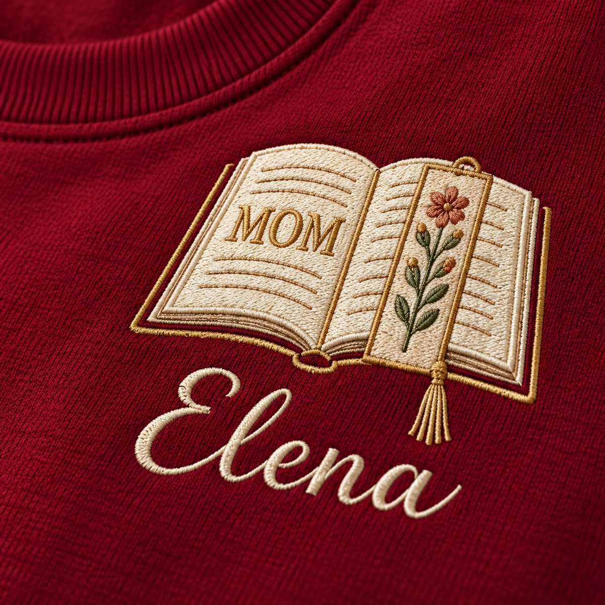 Motherhood Chapter sweatshirt concept mockup featuring an open book with a floral page and bookmark, sample personalization Elena, embroidery view
