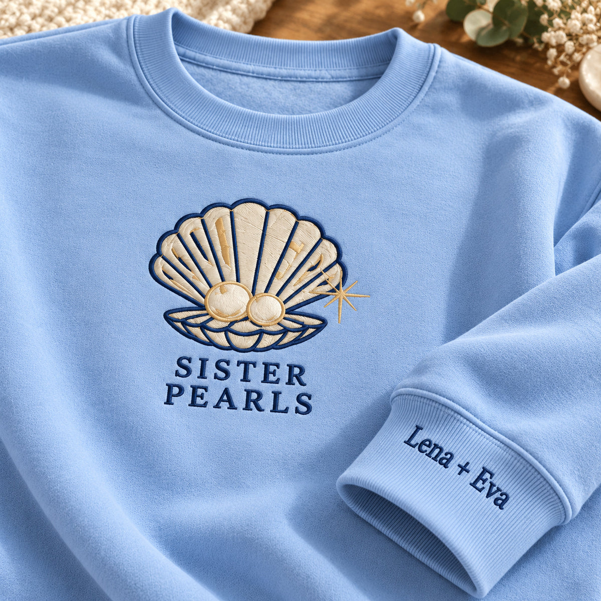 Sister Pearls sweatshirt concept mockup featuring an open seashell holding two pearls, sample personalization Lena + Eva, embroidery view